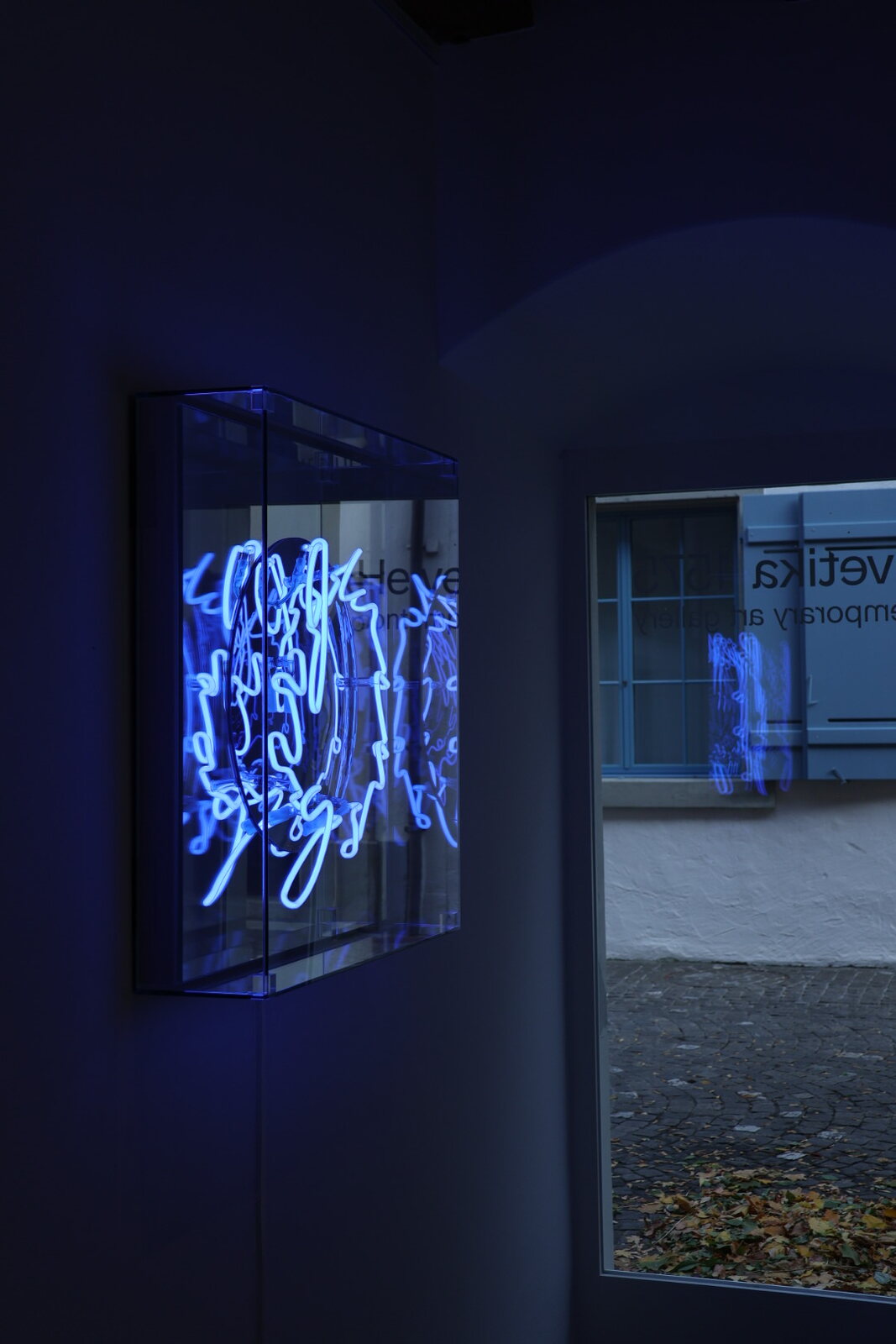 Blaue Neonskulptur in einer Glasbox, gespiegelt im Fenster zur Gasse