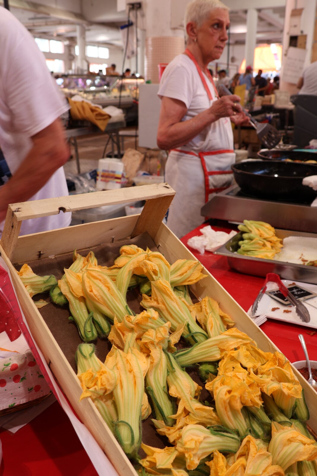 Kiste mit Zucchiniblüten, dahinter eine Köchin an der Pfanne
