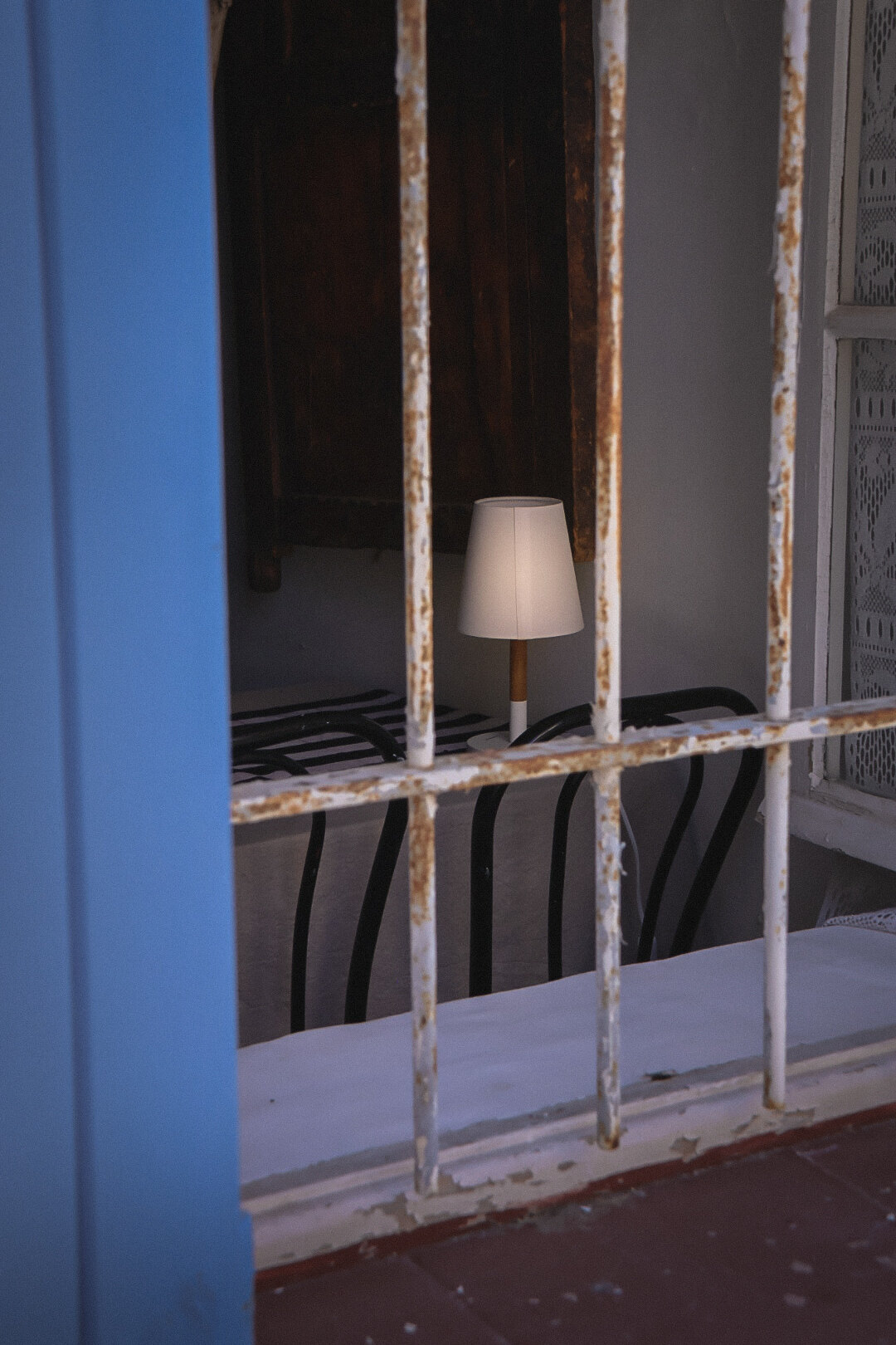 Blauer Fensterrahmen und rostiges weisses Gitter, dahinter eine kleine Lampe mit hellem Schirm